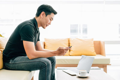 Asian male looking at the outside while using a mobile phone and working with laptop on the table. 