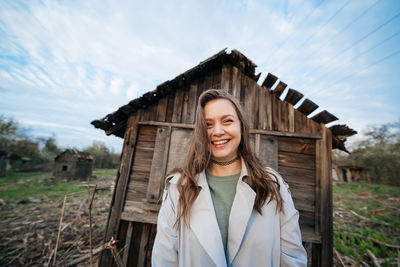 Beautiful girl with long hair in a grey trench coat next to an old wooden house outdoors in spring