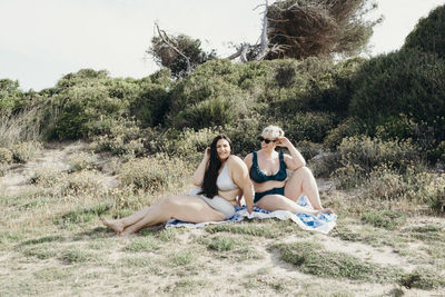 Delighted curvy friendly females in bikini sitting on blanket on shore and enjoying sunny day in summer