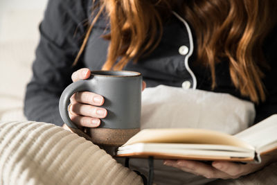 Midsection of woman holding mug while reading book on bed