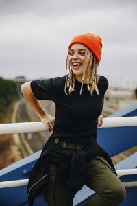 Cheerful young woman standing by railing on bridge against sky