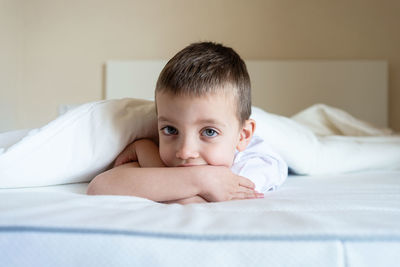 Adorable kid lying down on bed under blanket, while looking camera