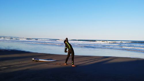 Full length of surfer standing at beach against clear blue sky