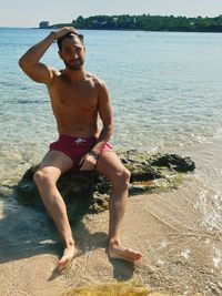 Portrait of shirtless man sitting on rock in sea