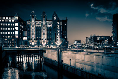 Illuminated bridge over river by buildings against sky at night