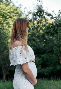 Pregnant woman wearing white dress, standing outdoor in orchard, holding pregnancy belly