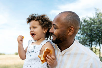 Positive loving african american father embracing adorable daughter eating tasty bun in summer field