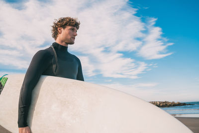 Young man carrying surfboard at beach against sky