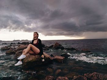 Portrait of young woman sitting on rock by sea against sky