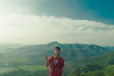 Portrait of man showing peace sign while standing on mountain against cloudy sky