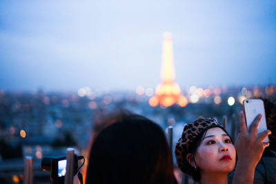 Portrait of woman photographing illuminated city against sky