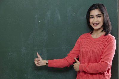 Portrait of happy female teacher gesturing thumbs up by blackboard