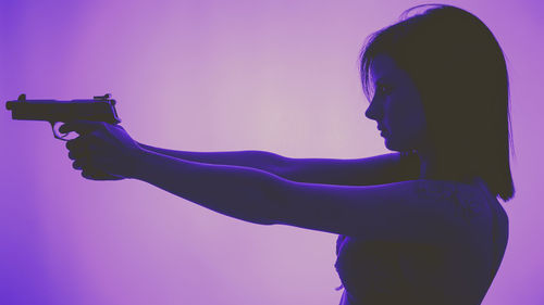 Side view of young woman holding handgun against purple background