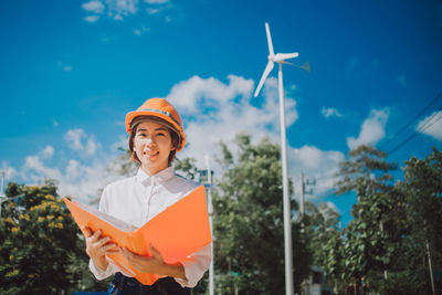 Portrait of female engineer in hardhat holding file while standing against windmill
