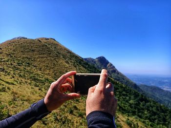 Cropped person photographing mountain with smart phone against clear blue sky
