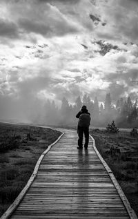 Rear view of man standing on boardwalk against cloudy sky