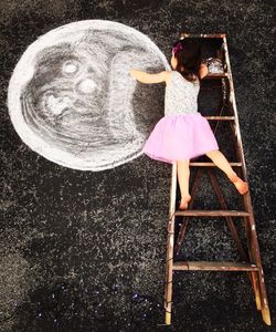 High angle view of girl lying on ladder while making chalk drawing on street