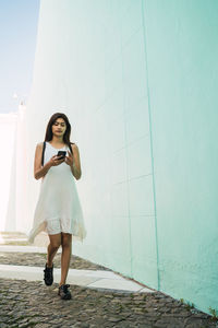 Full length of woman using phone walking against wall