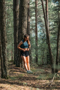 Young woman in sportswear leaned on a tree and straightens clothes in the forest during working out. 