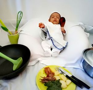 Baby in chef costume by food and kitchen utensils sleeping on bed at home