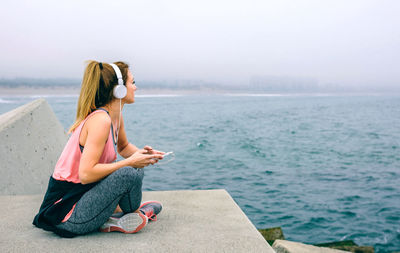 Young woman using mobile phone while sitting on sea against sky