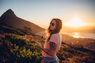 Young woman wearing sunglasses standing on land against sky during sunset