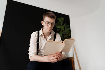 Low angle view of man reading book while sitting against wall
