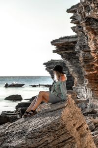 Side view of woman sitting on rock by sea against sky