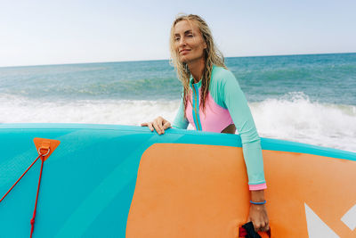 A young athletic blonde woman stands on the beach and holds a sup board.