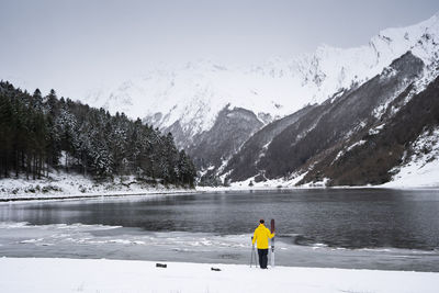 Man with ski at snow covered lakeshore in front of mountains