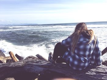 Rear view of woman sitting on driftwood at beach against sky