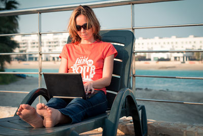 Woman working on laptop while siting on deck chair against railing