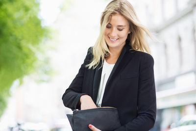 Smiling young woman searching something in her clutch bag outdoors