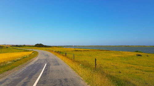 Empty road amidst field against clear blue sky