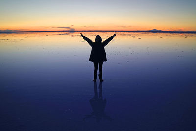 Silhouette of a happy traveler rising arms on mirror effect of uyuni salt flats at sunset, bolivia