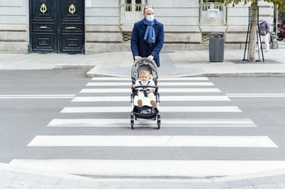 Man wearing face mask walking with baby stroller while crossing road in city