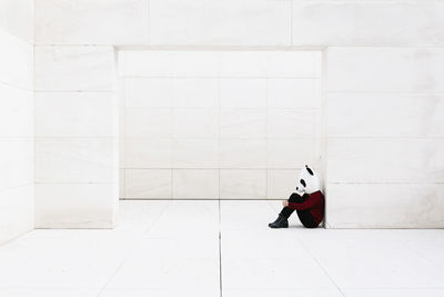 Depressed woman wearing panda mask sitting at doorway against white wall