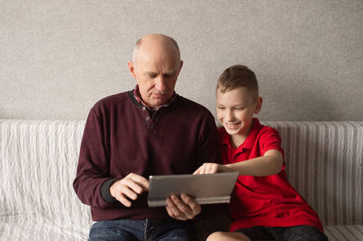 A boy teaches his grandfather how to use a tablet. the grandson is sitting next to the grandfather