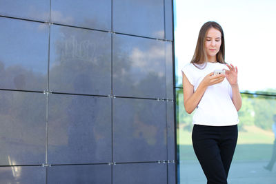 Young businesswoman using phone while standing against wall