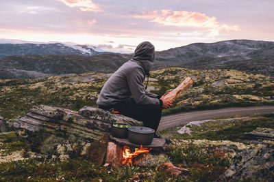 Full length of man sitting by fire against mountains and sky