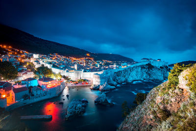 Illuminated city by sea against sky at dusk