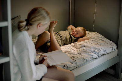 Sister reading picture book while sitting near brother with down syndrome lying on bed at home