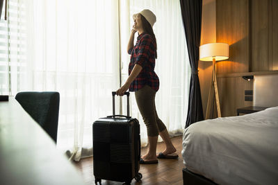 Full length of woman standing with suitcase by window