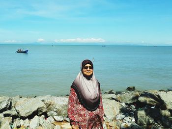 Woman in sunglasses and hijab standing by sea against sky