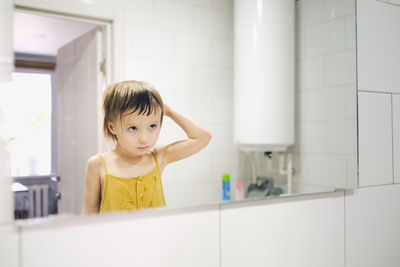 Child in yellow jumpsuit combs his own in front of mirror in bathroom