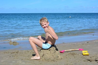 Full length portrait of shirtless boy gesturing while sitting at beach against sky