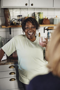 Happy mature woman with smoothie glass talking to female friend at home