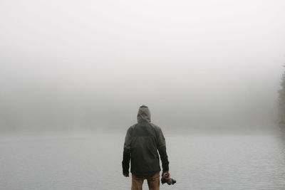 Rear view of man standing in front of lake in foggy weather