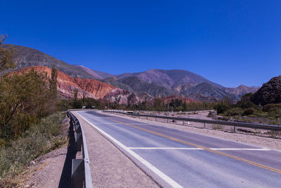 Empty road by mountains against clear blue sky