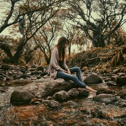 Young woman sitting on rock amidst stream flowing at forest